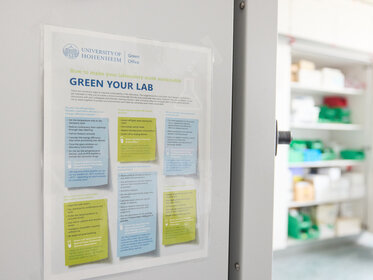 Foto: Ein Poster an einer Tür im Labor der Universität Hohenheim zeigt Tipps zur Verbesserung der Nachhaltigkeit im Labor. Der Text enthält Anweisungen zur Energieeinsparung und umweltfreundlichen Praktiken.