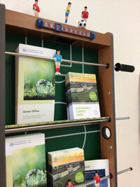 Foto: Ein ehemaliger Tischkicker, upgecycelt zu einem Wandregal, mit mehreren Broschüren der Universität Hohenheim, die auf dem Spielfeld liegen. Die Broschüren tragen Titel wie 'Sustainable Campus Guide' und 'Green Office'.