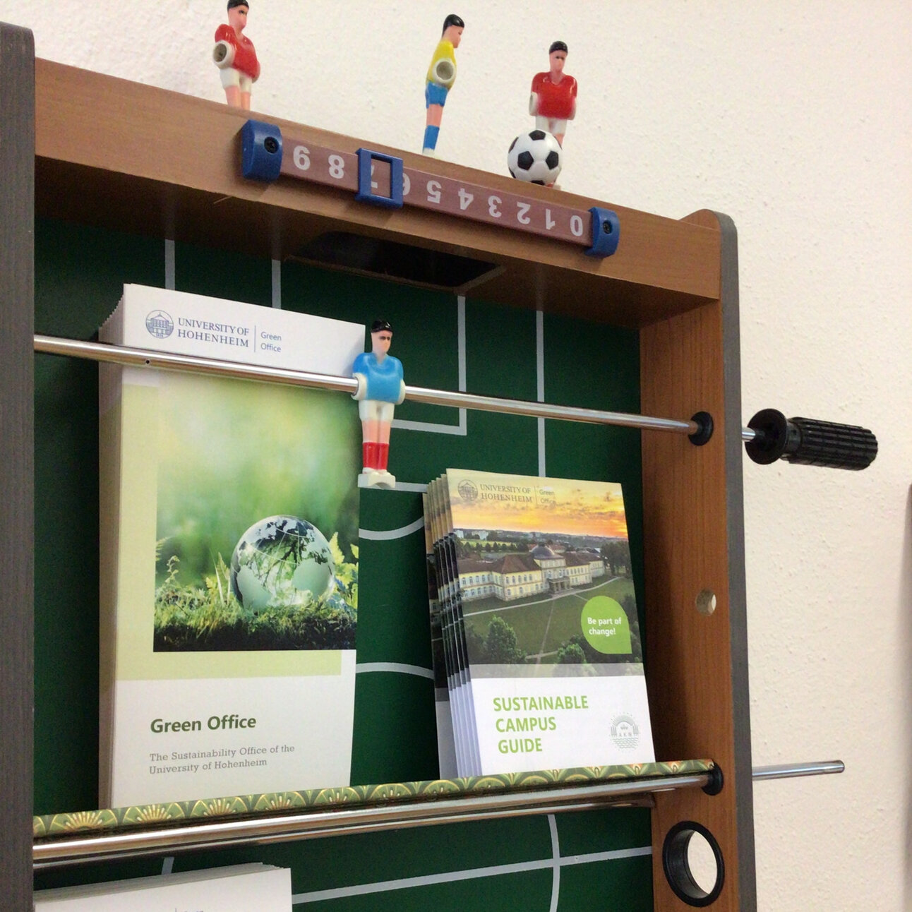 Foto: Ein ehemaliger Tischkicker, upgecycelt zu einem Wandregal, mit mehreren Broschüren der Universität Hohenheim, die auf dem Spielfeld liegen. Die Broschüren tragen Titel wie 'Sustainable Campus Guide' und 'Green Office'.