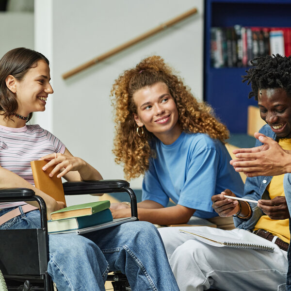 Foto: Eine Gruppe von Personen sitzt zusammen und unterhält sich. Eine Person sitzt in einem Rollstuhl und hält ein Buch. Im Hintergrund sind Bücherregale zu sehen.