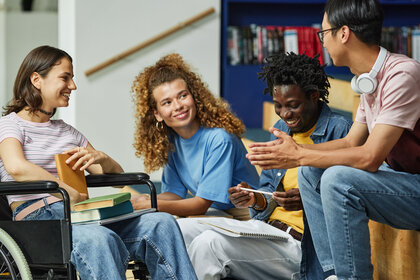 Foto: Eine Gruppe von Personen sitzt zusammen und unterhält sich. Eine Person sitzt in einem Rollstuhl und hält ein Buch. Im Hintergrund sind Bücherregale zu sehen.