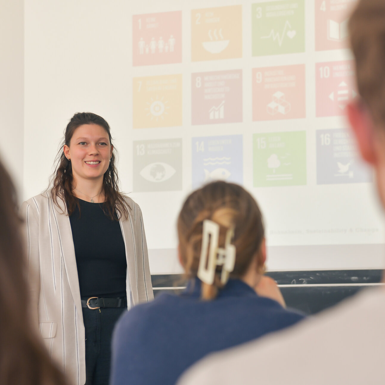 Foto: Eine Person steht vor einer Präsentation mit Symbolen der UN-Nachhaltigkeitsziele. Im Vordergrund sind unscharf Personen im Publikum zu sehen.
