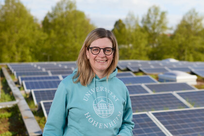 Foto: Eine Person in einem blauen Hoodie mit dem Logo der Universität Hohenheim steht vor einer Reihe von Solarpanelen. Im Hintergrund sind Bäume sichtbar.
