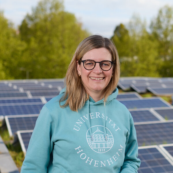 Foto: Eine Person in einem blauen Hoodie mit dem Logo der Universität Hohenheim steht vor einer Reihe von Solarpanelen. Im Hintergrund sind Bäume sichtbar.