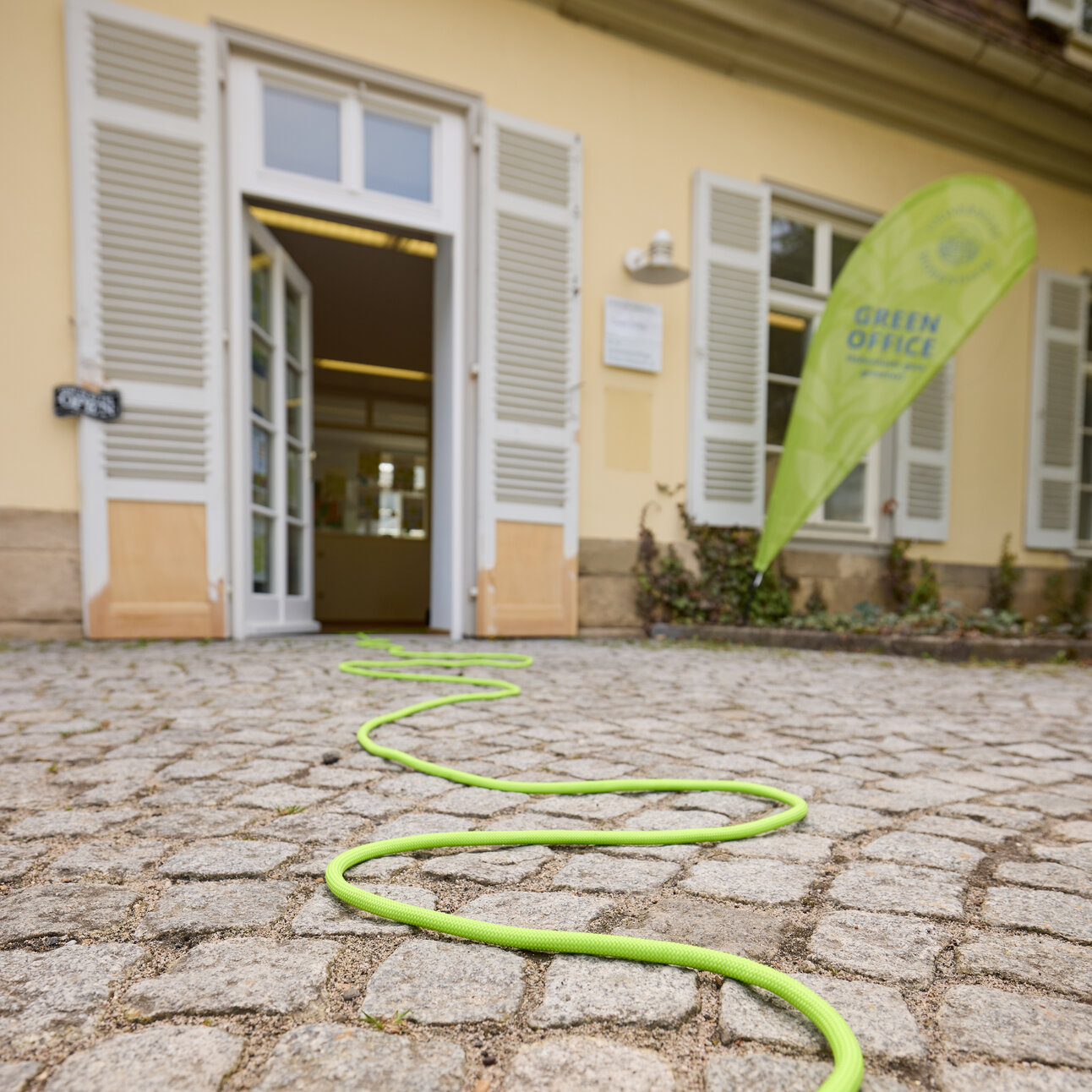 Foto: Ein grünes Kabel schlängelt sich über Pflastersteine zu einem Gebäude mit offenem Eingang. Ein Schild mit der Aufschrift 'Green Office' ist sichtbar.