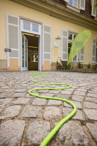 Foto: Ein grünes Kabel schlängelt sich über Pflastersteine zu einem Gebäude mit offenem Eingang. Ein Schild mit der Aufschrift 'Green Office' ist sichtbar.