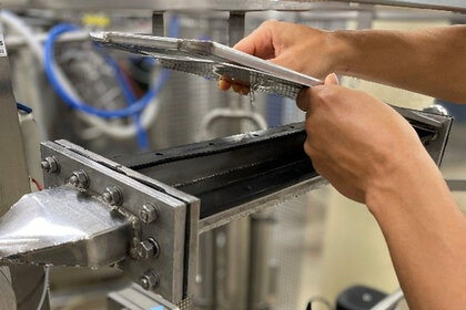 Foto: Eine Person arbeitet an einer industriellen Maschine mit mehreren Metallrohren und Ventilen. Die Person hält ein rechteckiges Metallteil in den Händen, während sie an der Maschine arbeitet.