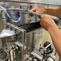 Foto: Eine Person arbeitet an einer industriellen Maschine mit mehreren Metallrohren und Ventilen. Die Person hält ein rechteckiges Metallteil in den Händen, während sie an der Maschine arbeitet.
