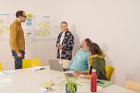 Foto: Vier Personen in einem Büro bei einem Meeting. Eine Person steht vor einem Whiteboard mit Notizen und spricht zu den anderen, die an einem Tisch mit Laptops und Notizbüchern sitzen.