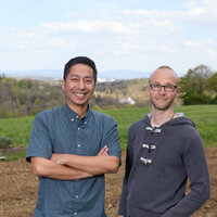 Foto: Zwei Personen stehen auf einem Feld mit Blick auf eine hügelige Landschaft im Hintergrund. Der Himmel ist leicht bewölkt und es gibt einige Bäume und Gebäude in der Ferne.