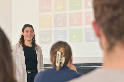 Foto: Eine Person steht vor einer Präsentation mit Symbolen der UN-Nachhaltigkeitsziele. Im Vordergrund sind unscharf Personen im Publikum zu sehen.