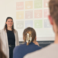 Foto: Eine Person steht vor einer Präsentation mit Symbolen der UN-Nachhaltigkeitsziele. Im Vordergrund sind unscharf Personen im Publikum zu sehen.