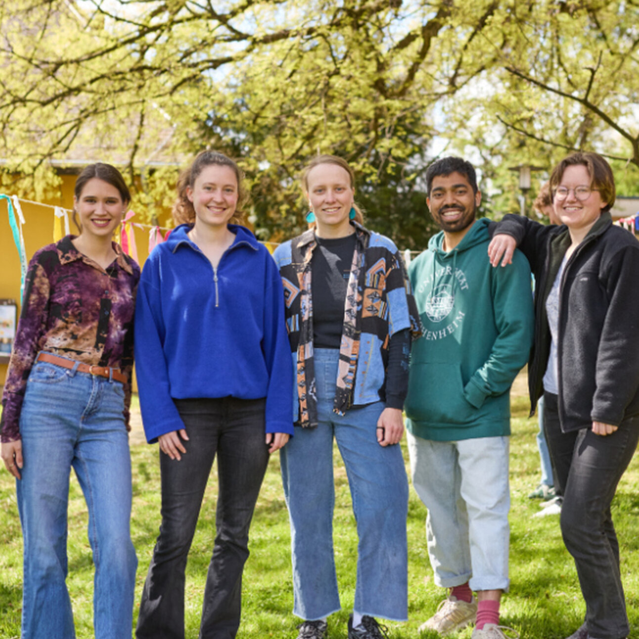 Foto: Fünf Personen stehen auf einer Wiese vor einem Gebäude mit bunten Bändern im Hintergrund. Die Szene ist von Bäumen umgeben und vermittelt eine entspannte Atmosphäre.