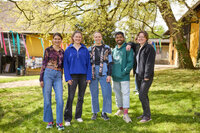 Foto: Fünf Personen stehen auf einer Wiese vor einem Gebäude mit bunten Bändern im Hintergrund. Die Szene ist von Bäumen umgeben und vermittelt eine entspannte Atmosphäre.