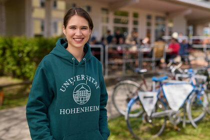 Foto: Eine Person trägt einen grünen Hoodie mit dem Logo der Universität Hohenheim. Im Hintergrund sind Fahrräder und eine Terrasse mit Menschen zu sehen.