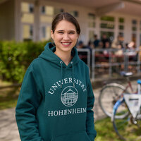 Foto: Eine Person trägt einen grünen Hoodie mit dem Logo der Universität Hohenheim. Im Hintergrund sind Fahrräder und eine Terrasse mit Menschen zu sehen.