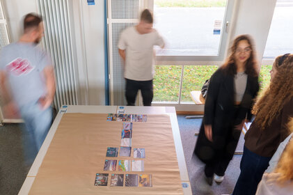 Foto: Mehrere Personen stehen um einen Tisch in einem Raum. Auf dem Tisch liegen Karten in einer Anordnung. Der Raum hat große Fenster, durch die Tageslicht einfällt.