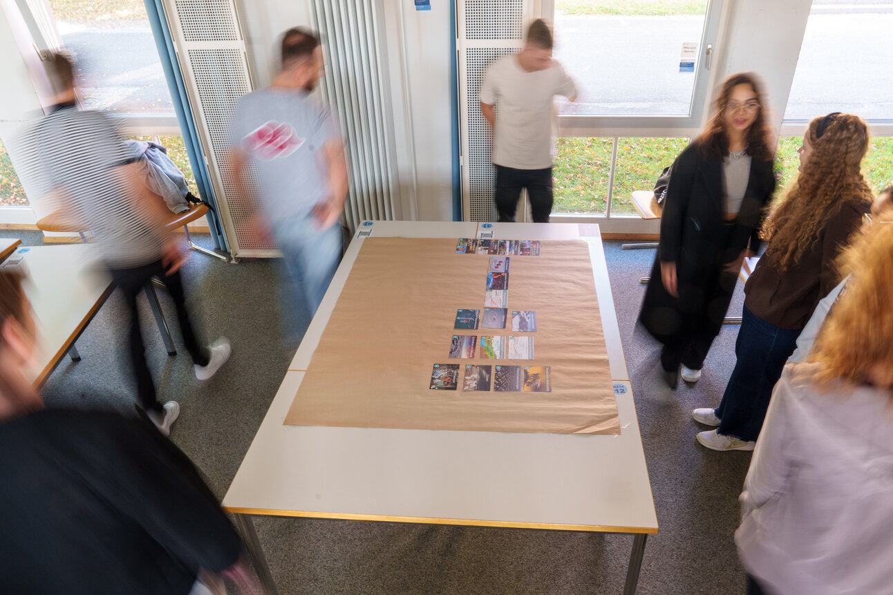 Foto: Mehrere Personen stehen um einen Tisch in einem Raum. Auf dem Tisch liegen Karten in einer Anordnung. Der Raum hat große Fenster, durch die Tageslicht einfällt.