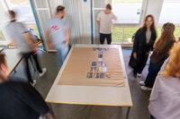 Foto: Mehrere Personen stehen um einen Tisch in einem Raum. Auf dem Tisch liegen Karten in einer Anordnung. Der Raum hat große Fenster, durch die Tageslicht einfällt.