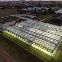 Foto: Luftaufnahme eines großen, beleuchteten Gewächshauses bei Dämmerung. Das Gebäude ist von landwirtschaftlichen Flächen umgeben, mit mehreren kleineren Gebäuden im Hintergrund.