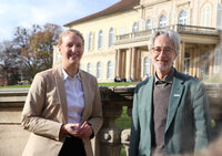 Foto: Zwei Personen stehen vor Schloss Hohenheim. Die Personen tragen formelle Kleidung. Im Hintergrund ist das historische Gebäude mit symmetrischem Design und Säulen zu sehen.