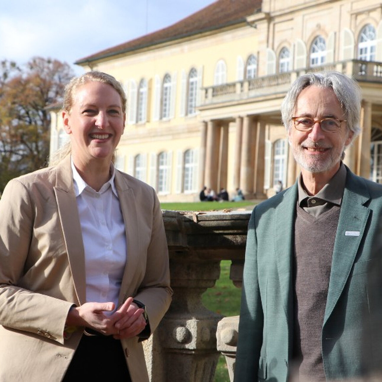 Foto: Zwei Personen stehen vor Schloss Hohenheim. Die Personen tragen formelle Kleidung. Im Hintergrund ist das historische Gebäude mit symmetrischem Design und Säulen zu sehen.