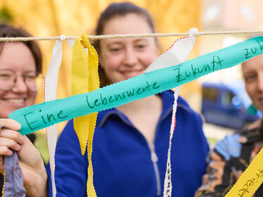 Foto: Drei Personen halten bunte Bänder mit handgeschriebenen Botschaften wie 'Eine lebenswerte Zukunft'. Die Bänder sind an einer Schnur befestigt und im Freien aufgenommen.