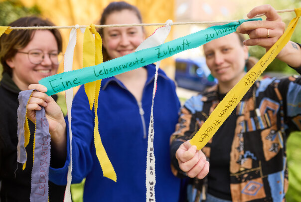 Foto: Drei Personen halten bunte Bänder mit handgeschriebenen Botschaften wie 'Eine lebenswerte Zukunft'. Die Bänder sind an einer Schnur befestigt und im Freien aufgenommen.