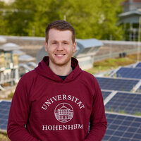 Foto: Eine Person in einem roten Hoodie mit dem Logo der Universität Hohenheim steht auf einem Dach mit Solarpanelen. Im Hintergrund sind grüne Bäume sichtbar.