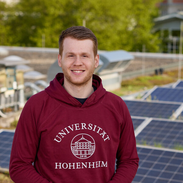 Foto: Eine Person in einem roten Hoodie mit dem Logo der Universität Hohenheim steht auf einem Dach mit Solarpanelen. Im Hintergrund sind grüne Bäume sichtbar.