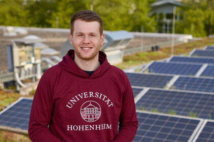 Foto: Eine Person in einem roten Hoodie mit dem Logo der Universität Hohenheim steht auf einem Dach mit Solarpanelen. Im Hintergrund sind grüne Bäume sichtbar.