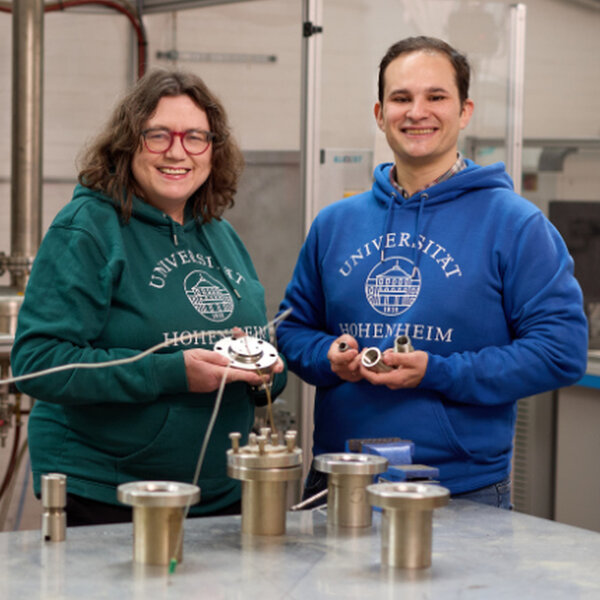 Foto: Zwei Personen in Pullovern der Universität Hohenheim stehen in einem Labor. Sie halten metallische Geräte in den Händen. Im Hintergrund sind industrielle Apparaturen sichtbar.