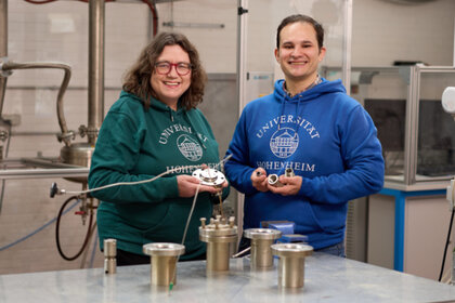 Foto: Zwei Personen in Pullovern der Universität Hohenheim stehen in einem Labor. Sie halten metallische Geräte in den Händen. Im Hintergrund sind industrielle Apparaturen sichtbar.
