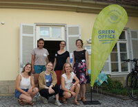 Foto: Eine Gruppe von Personen steht vor einem Gebäude mit einem Banner der Universität Hohenheim, das 'GREEN OFFICE Hohenheim goes green(er)' zeigt.