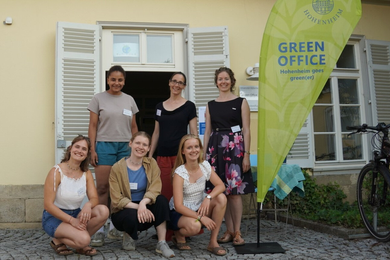 Foto: Eine Gruppe von Personen steht vor einem Gebäude mit einem Banner der Universität Hohenheim, das 'GREEN OFFICE Hohenheim goes green(er)' zeigt.