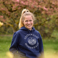Foto: Eine Person steht auf einem Weg in einem Park und trägt einen blauen Hoodie mit dem Logo der Universität Hohenheim. Im Hintergrund sind blühende Bäume und ein gepflasterter Weg zu sehen.