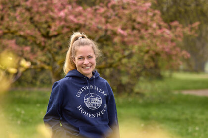 Foto: Eine Person steht auf einem Weg in einem Park und trägt einen blauen Hoodie mit dem Logo der Universität Hohenheim. Im Hintergrund sind blühende Bäume und ein gepflasterter Weg zu sehen.