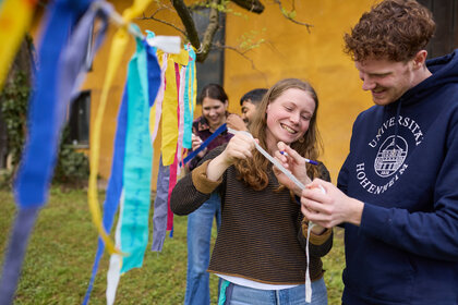 Foto: Mehrere Personen stehen im Freien vor einem gelben Gebäude. Sie halten bunte Stoffstreifen und schreiben darauf. Eine Person trägt einen Pullover mit dem Logo der Universität Hohenheim.