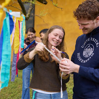 Foto: Mehrere Personen stehen im Freien vor einem gelben Gebäude. Sie halten bunte Stoffstreifen und schreiben darauf. Eine Person trägt einen Pullover mit dem Logo der Universität Hohenheim.