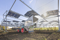 Foto: Ein Traktor mit landwirtschaftlichem Gerät steht unter einer Struktur aus Solarpanelen auf einem Feld. Die Solarpanele sind in Reihen angeordnet und die Sonne scheint durch sie hindurch.
