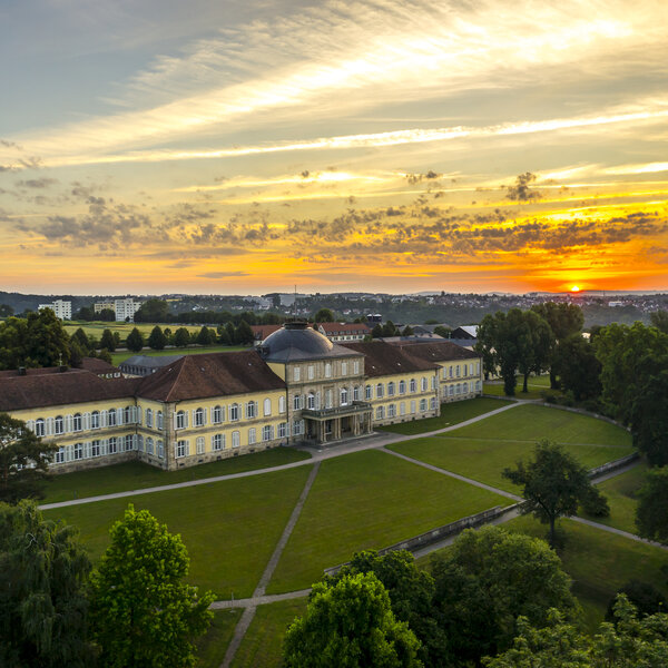 Foto: Schloss Hohenheim bei Sonnenuntergang, umgeben von gepflegten Rasenflächen und Bäumen. Der Himmel zeigt lebendige Farben von Orange und Gelb.