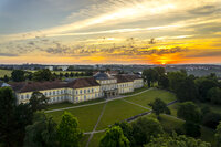Foto: Schloss Hohenheim bei Sonnenuntergang, umgeben von gepflegten Rasenflächen und Bäumen. Der Himmel zeigt lebendige Farben von Orange und Gelb.