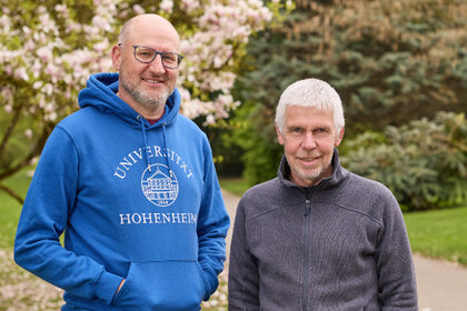 Foto: Zwei Personen stehen im Freien im Park der Universität Hohenheim. Eine Person trägt einen blauen Hoodie mit der Aufschrift 'Universität Hohenheim'. Im Hintergrund sind blühende Bäume und ein gepflegter Rasen zu sehen.