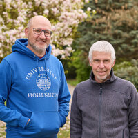 Foto: Zwei Personen stehen im Freien im Park der Universität Hohenheim. Eine Person trägt einen blauen Hoodie mit der Aufschrift 'Universität Hohenheim'. Im Hintergrund sind blühende Bäume und ein gepflegter Rasen zu sehen.