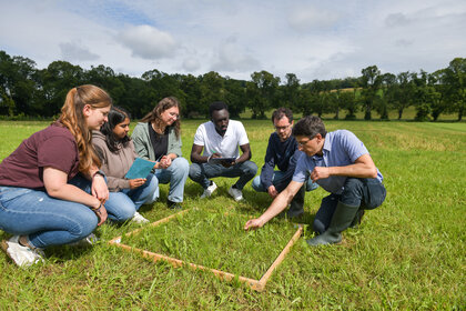 Foto: Eine Gruppe von Personen kniet auf einer Wiese um einen quadratischen Holzrahmen. Sie beobachten den Boden und notieren sich Details. Im Hintergrund sind Bäume und ein bewölkter Himmel zu sehen.