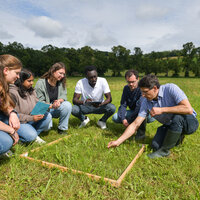 Foto: Eine Gruppe von Personen kniet auf einer Wiese um einen quadratischen Holzrahmen. Sie beobachten den Boden und notieren sich Details. Im Hintergrund sind Bäume und ein bewölkter Himmel zu sehen.