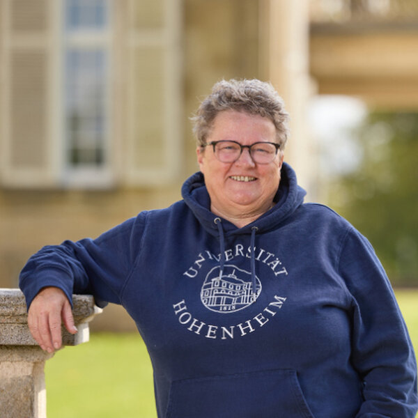 Foto: Eine Person steht draußen und lehnt an einer Mauer. Sie trägt einen blauen Hoodie mit dem Aufdruck 'Universität Hohenheim'. Im Hintergrund ist ein Gebäude und Grünfläche sichtbar.