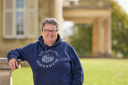 Foto: Eine Person steht draußen und lehnt an einer Mauer. Sie trägt einen blauen Hoodie mit dem Aufdruck 'Universität Hohenheim'. Im Hintergrund ist ein Gebäude und Grünfläche sichtbar.