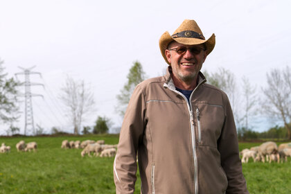 Foto: Eine Person mit Hut steht auf einer grünen Weide, auf der Schafe grasen. Im Hintergrund sind Bäume und ein Strommast zu sehen.