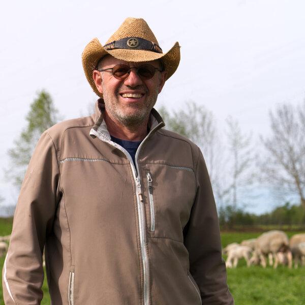 Foto: Eine Person mit Hut steht auf einer grünen Weide, auf der Schafe grasen. Im Hintergrund sind Bäume und ein Strommast zu sehen.